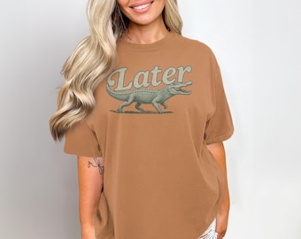 Peut inclure: Un t-shirt marron avec le mot "Later" au-dessus d'une illustration d'alligator vert. Le t-shirt a des manches courtes et une coupe décontractée. Le design est d'inspiration vintage.