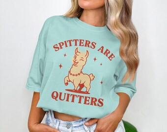 以下が含まれることがあります： 赤色の「Spitters Are Quitters」の文字が入った水色のTシャツ。花のネックレスをつけた漫画のラマが描かれています。ラマは後ろ足で立ってウィンクをしています。カジュアルな普段着アイテムです。