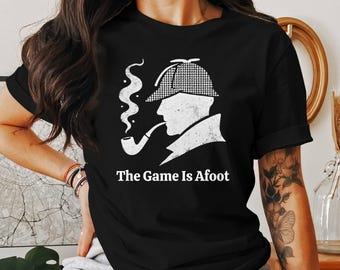 May include: Black t-shirt featuring a white graphic of a silhouette of a person with a pipe and a checkered hat, with the text "The Game Is Afoot" in white below the graphic. The shirt is a classic crew neck.
