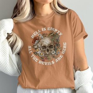May include: A tan t-shirt featuring a circular graphic with a skull surrounded by flowers and the text "HELL IS EMPTY ALL THE DEVILS ARE HERE" in white. The shirt has short sleeves and a crew neck.