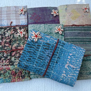 Op de afbeelding: Een verzameling handgemaakte stoffen tijdschriften in verschillende kleuren en patronen. Elk tijdschrift is versierd met een decoratieve bloem en gebonden met een rood koord. De tijdschriften hebben een mix van bloemen, geometrische en abstracte ontwerpen.