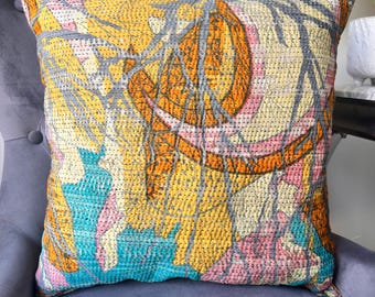 Op de afbeelding: Vierkant decoratief kussen met een kleurrijk patchwork ontwerp. Het kussen heeft een levendige mix van oranje, geel, turquoise, roze en grijstinten. De stof heeft een gewatteerde textuur. Het kussen ligt op een grijze fauteuil.