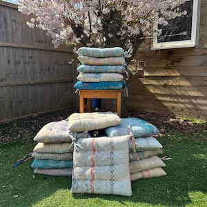 May include: A stack of colorful, patterned cushions made from recycled saris. The cushions are arranged in a pyramid shape on a wooden table. The cushions are perfect for adding a touch of bohemian style to any room.