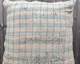 Puede incluir: Un cojín cuadrado con una base de tela texturizada en color blanco roto y un patrón de cuadrícula en azul verdoso y azul claro. La superficie muestra detalles de costura visibles y pequeñas manchas de color azul oscuro dispersas.