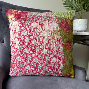 May include: A square cushion with a patchwork design. It features a vibrant mix of floral patterns in red, pink, green, and white. The cushion is placed on a grey upholstered chair, highlighting its colours and textures.