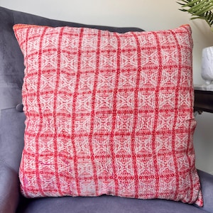 Puede incluir: Un cojín decorativo cuadrado con un patrón geométrico rojo y blanco. El cojín está hecho de una tela texturizada y está colocado en un sillón gris. El patrón consiste en una cuadrícula de cuadrados con un diseño repetitivo.