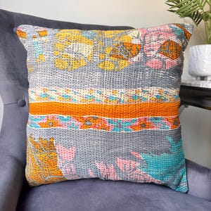 May include: Square cushion with a patchwork design. The cushion features a grey background with floral and geometric patterns in orange, yellow, pink, and teal. The cushion is placed on a grey armchair.