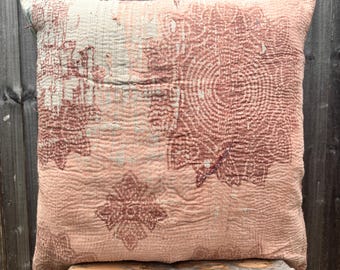 Op de afbeelding: Een vierkant sierkussen van licht perzikkleurige en oudroze stof met een zichtbaar stikselpatroon in kantha-stijl. Het textiel vertoont een vervaagde bloemen- en geometrische print in donkerdere rozetinten, met gerafelde randen aan de bovenkant.