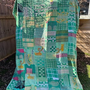 Puede incluir: Una colcha de retazos hecha de varias telas verdes, amarillas y rosas. La colcha está hecha de muchos cuadrados y rectángulos diferentes, cada uno con un patrón único. La colcha está doblada y colocada sobre una superficie cubierta de hierba.