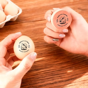May include: A close-up of a brown egg stamped with "SMITH FARM FRESH EGGS" in black ink. The stamp is held in a person's hand, with a carton of eggs in the background. The stamp features a graphic of a chicken and eggs.