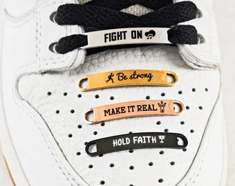 May include: Close-up of a white sneaker with black laces and decorative metal shoelace tags. The tags are gold, copper, and black, with motivational phrases like "FIGHT ON," "Be strong," "MAKE IT REAL," and "HOLD FAITH."