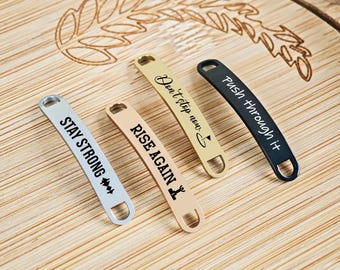 Puede incluir: Cuatro etiquetas metálicas para zapatos con frases motivadoras: "Stay Strong", "Rise Again", "Don't stop now" y "Push through it". Las etiquetas son plateadas, beige, doradas y negras, con texto negro. Tienen un lazo.