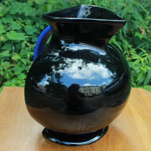 May include: A glossy black pitcher with a blue handle. The pitcher has a rounded body and a flared rim. The surface reflects the sky and surrounding environment. The pitcher sits on a wooden surface.