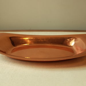 Puede incluir: Una fuente oblonga de color cobre, con una forma suavemente curvada. La fuente tiene una superficie lisa y reflectante, que capta la luz y crea reflejos. Los bordes están ligeramente elevados y el diseño general es simple y elegante. Esta fuente decorativa es adecuada para servir o exhibir.
