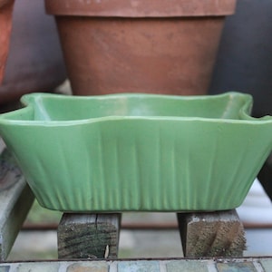 May include: A green ceramic planter with a fluted design. The planter is empty and has a square shape.