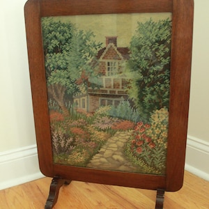 Op de afbeelding: Een ingelijste tapisserie met een huis omgeven door een tuin. De tapisserie is geplaatst in een donkerhouten frame met decoratieve poten. De scène toont een bakstenen huis, een stenen pad en kleurrijke bloemen.