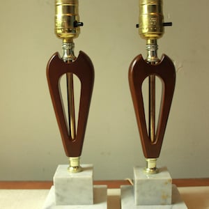 Może przedstawiać: Para lamp stołowych w stylu vintage. Każda lampa ma ciemne drewno w kształcie łzy z mosiężnym prętem środkowym i akcentami. Lampy stoją na białych marmurowych podstawach i mają mosiężne oprawki żarówek.