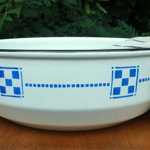May include: A white enamelware bowl with a black rim and blue checkered and dotted design. A striped towel hangs over the side of the bowl. The bowl is sitting on a wooden surface.