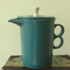 May include: A teal ceramic French press coffee maker with a lid and a white knob. The coffee maker has a spout and two loop handles. It is sitting on top of a stack of old books with gold lettering.