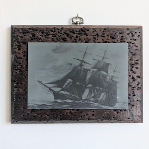May include: A rectangular wall hanging featuring a dark, textured, porous wooden frame with a metal hanging loop at the top. The center contains a monochromatic print of a sailing ship with multiple masts and dark sails navigating choppy waters.