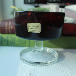 May include: A dark red glass goblet with a wide bowl and a clear stem and base. The goblet has a sticker that reads "luminarc, VERRE D'ARQUES, FRANCE". The goblet is in front of two books.