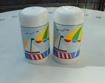 May include: Two white ceramic salt and pepper shakers. Each shaker features a beach scene with a sailboat, yellow and green umbrella, and a seagull against a light blue background. The base has red, white, and blue stripes.