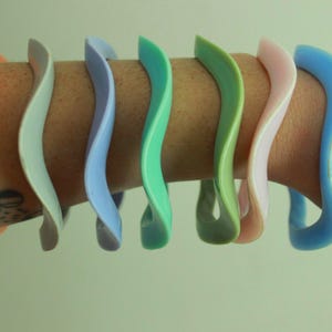 Könnte beinhalten: Eine Sammlung farbenfroher, wellenförmiger Armreifen in Orange, Grau, Blau, Grün, Rosa und Hellblau. Die Armreifen werden an einem Arm getragen und zeigen ihr einzigartiges, geschwungenes Design. Jeder Armreif hat eine glatte, glänzende Oberfläche.