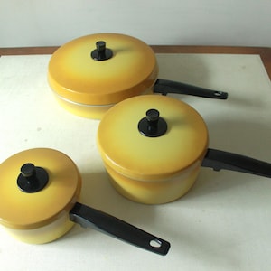 Puede incluir: Conjunto de tres utensilios de cocina amarillos y negros con tapas. Las ollas están graduadas en tamaño y tienen asas negras.