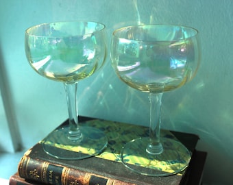 May include: Two iridescent champagne glasses with long stems. The glasses sit on a stack of antique books with gold lettering. The glasses have a pearlescent sheen, reflecting shades of green, blue, and yellow.