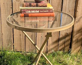 Op de afbeelding: Een ronde bijzettafel met een glazen blad en een goudkleurig metalen frame met gekruiste poten. Een terracotta pot met een groene plant staat op een stapel van drie gebonden boeken, waaronder een boek met de titel Your Isadora.