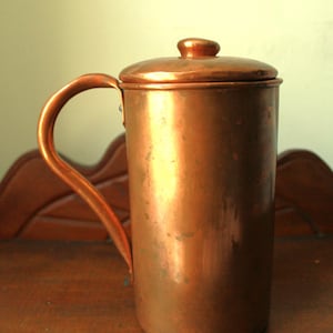 May include: A copper-colored, cylindrical mug with a lid and handle. The mug has a rounded handle and a small knob on the lid. The copper has a warm, reflective sheen, suggesting it is made of solid copper. The mug is likely used for beverages.