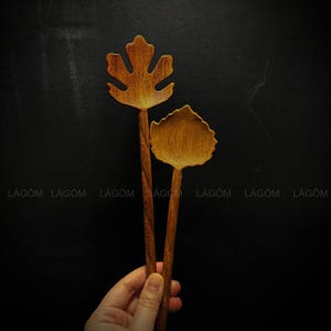 Op de afbeelding: Twee houten lepels met blaadjesvormige kommen. De lepels zijn bruin en hebben een natuurlijke houten afwerking. De lepels worden in een hand tegen een zwarte achtergrond gehouden.