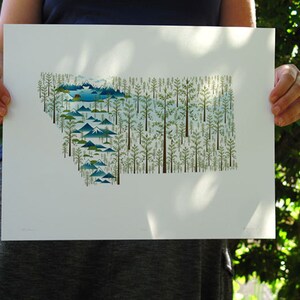 May include: A white print of the state of Montana, outlined in green trees and blue mountains. The print is held by a person's hands.