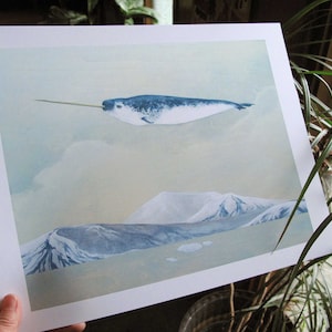 May include: A watercolor painting of a narwhal flying over a snowy mountain range. The narwhal is blue and white and has a long, spiraled tusk. The mountains are a light blue and white, and the sky is a light blue.