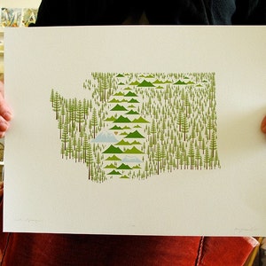 May include: A print of the state of Washington, USA, made up of green trees and mountains. The state outline is filled with small green trees and the mountains are represented by green shapes. The print is on white paper and is being held by a person's hands.