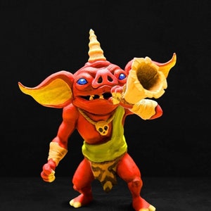 May include: A red and yellow goblin figurine with a horn on its head and a yellow trumpet in its hand. The goblin is wearing a green loincloth and a necklace with a skull pendant.