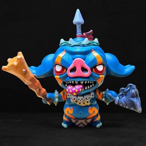 May include: A blue and orange painted figurine of a pig-like creature with a spear on its head, holding a wooden club in one hand and a blue boot in the other. The creature has a pink snout, red eyes, and a pink tongue with white polka dots.