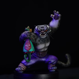 May include: A snow leopard figurine with a black and white fur pattern, holding a green and purple fabric with a red and gold design. The figurine is standing on a black base.