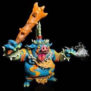 May include: A blue and orange painted figurine of a boar-like creature with a crown of leaves and a club in one hand. The creature has a large, open mouth with a pink tongue and a fierce expression.