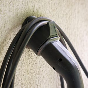 Peut inclure: Gros plan sur un connecteur et un câble de recharge de véhicule électrique noirs. Le connecteur est branché sur un mur, le câble étant enroulé autour. Le mur a une surface beige clair texturée.
