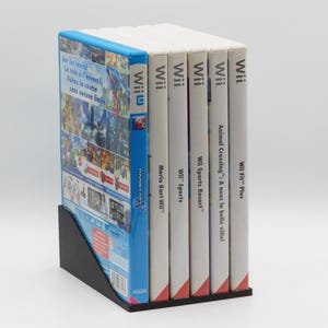 Peut inclure: Un ensemble de six boîtiers de jeux vidéo pour consoles Wii et Wii U, disposés verticalement dans un support en plastique noir. Le boîtier avant est bleu, tandis que les autres sont blancs avec du texte noir identifiant des titres comme Mario Kart.