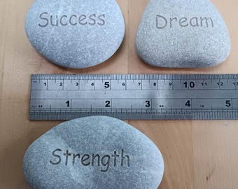 Puede incluir: Tres piedras grises con las palabras "Éxito", "Sueño" y "Fuerza" grabadas en ellas. Las piedras están dispuestas en una fila sobre una superficie de madera con una regla debajo.
