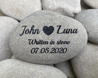 May include: A smooth, light gray river stone engraved with the black text John, a heart symbol, and Luna. Below the names, the text reads Written in stone followed by the date 07.05.2020. The stone has a natural, rounded, pebble-like shape.