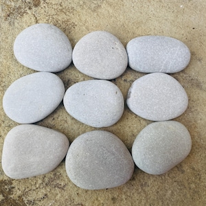 Può includere: Dieci ciottoli di fiume lisci di colore grigio chiaro, disposti a griglia su una superficie beige.