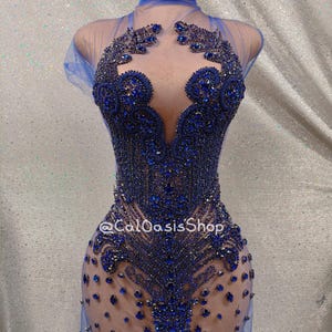 Pode incluir: Um vestido azul transparente de mini saia com gola alta e um corpete justo. O vestido é coberto de pedrarias azuis e tem uma saia transparente. O vestido está em um manequim e o texto "@CalOasisShop" é visível no vestido.