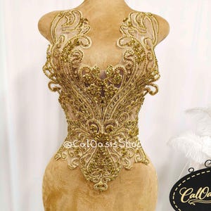 May include: A gold-colored bodice with intricate embroidery and beadwork, displayed on a mannequin. The design features swirling patterns and a deep V-neck, creating an elegant and ornate look. The bodice is likely for formal wear.