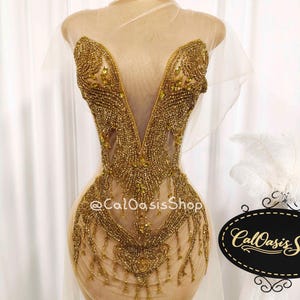 May include: A gold beaded dress on a mannequin. The dress has a deep V-neck and detailed beadwork. The dress is decorated with gold beads and hanging embellishments. The dress is displayed on a mannequin with a sheer overlay.