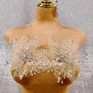 May include: A tan mannequin bust displays two elaborate, floral-shaped body adornments. The silver-toned pieces are embellished with clear crystals and delicate wire detailing. The text "@CalOasisShop" is visible on the bust.