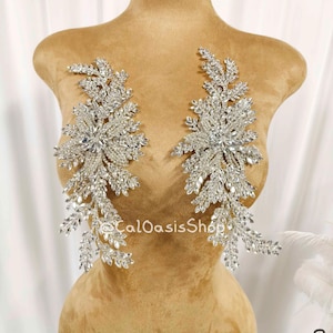 May include: Two silver rhinestone appliques in a floral design. Each applique features a central cluster of large rhinestones surrounded by smaller stones and leaf-like accents. The appliques are displayed on a mannequin.