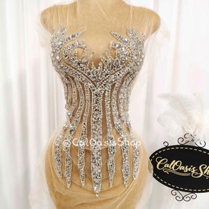 May include: A silver and clear rhinestone embellished bodice on a mannequin. The bodice is made of a sheer, nude-colored fabric and is adorned with intricate patterns of sparkling rhinestones. The design includes vertical lines and floral-like accents.